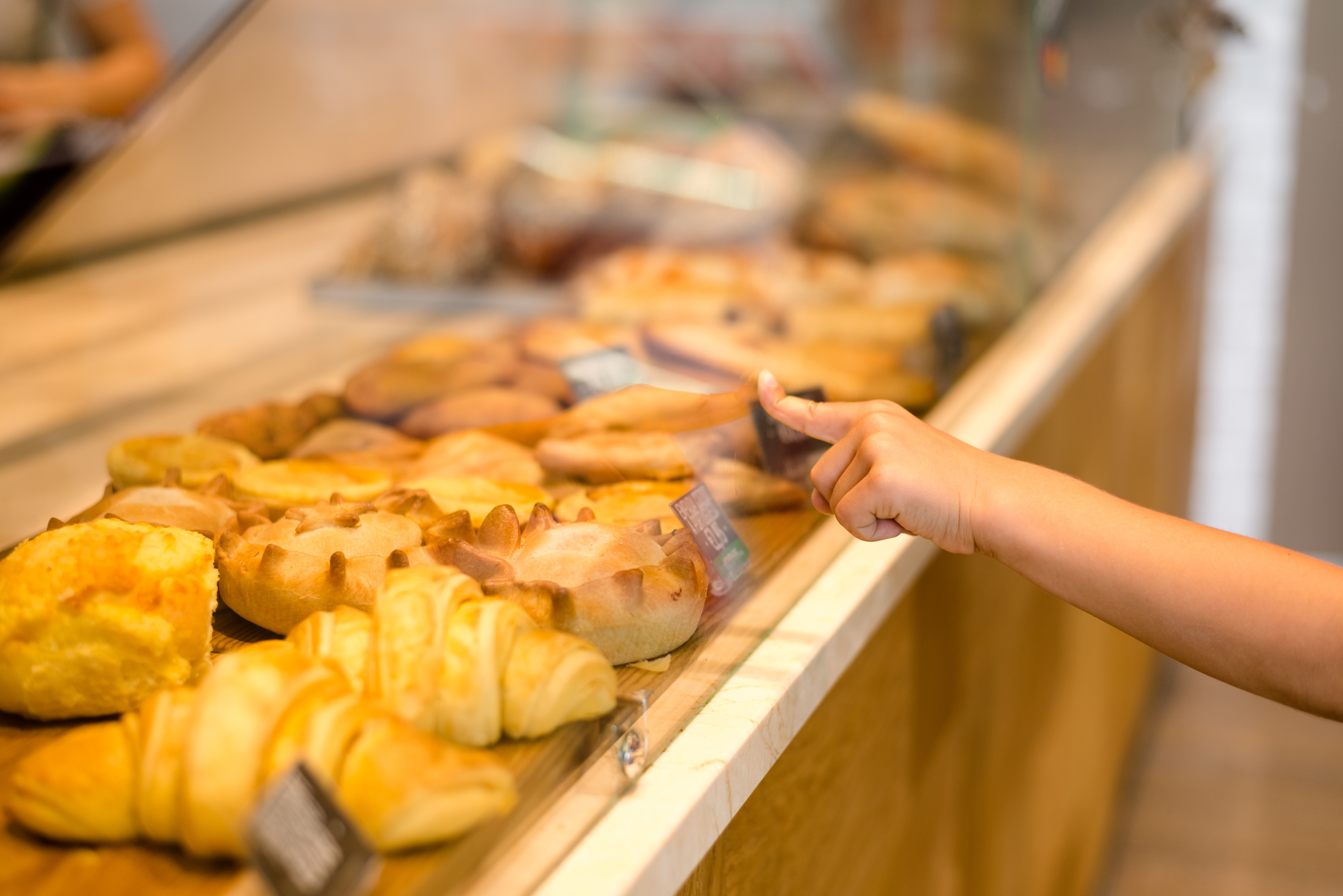 Picture of tasty pastry on store glass showcase. Closeup of child's hand with index finger pointing on some cake on blurred indoor background.