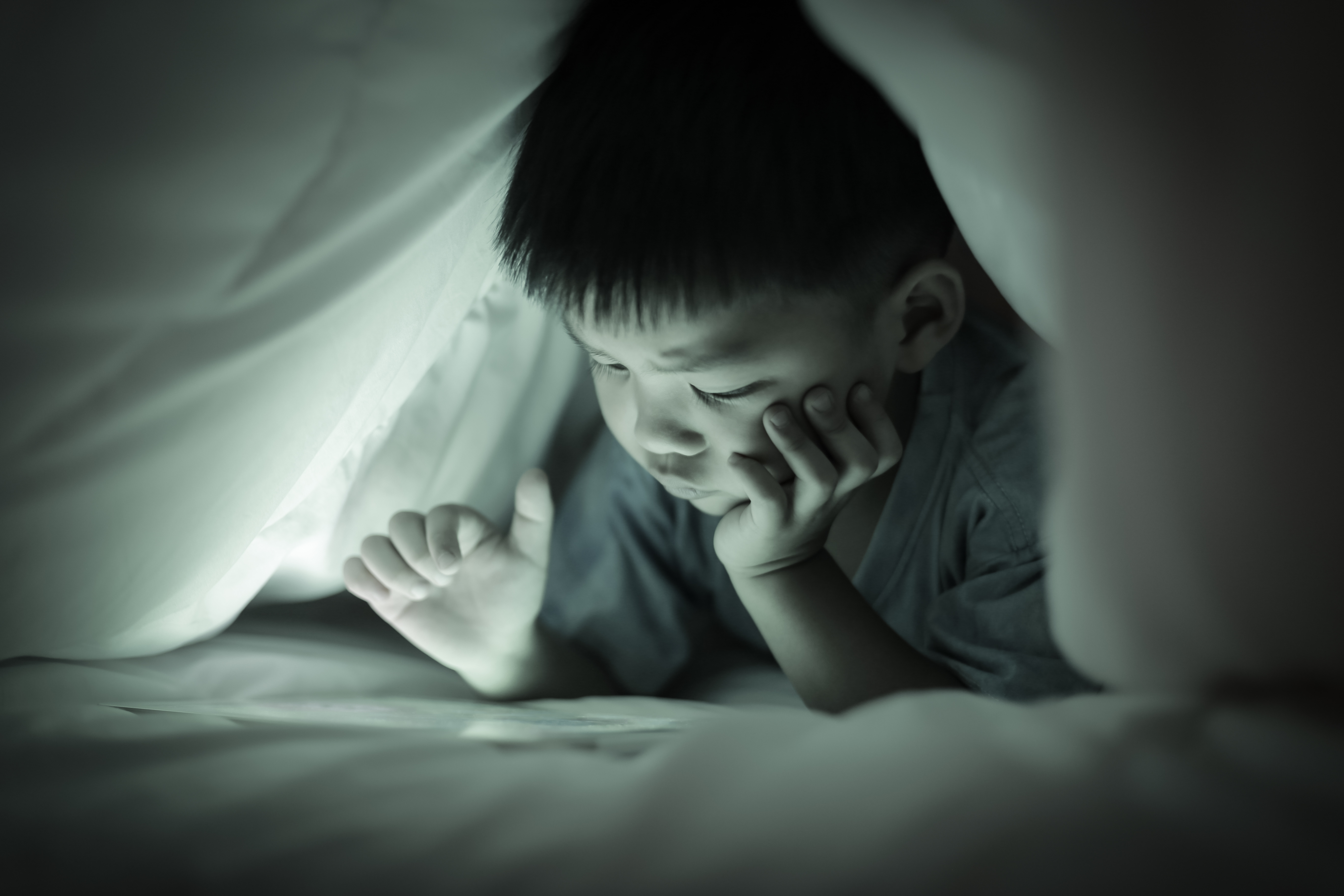 Little boy watching online tablet while lying on bed under white duvet, in the bedroom at night, bright screen light reflex on little child face and his finger hand pointing at  screen. Sepia tone.