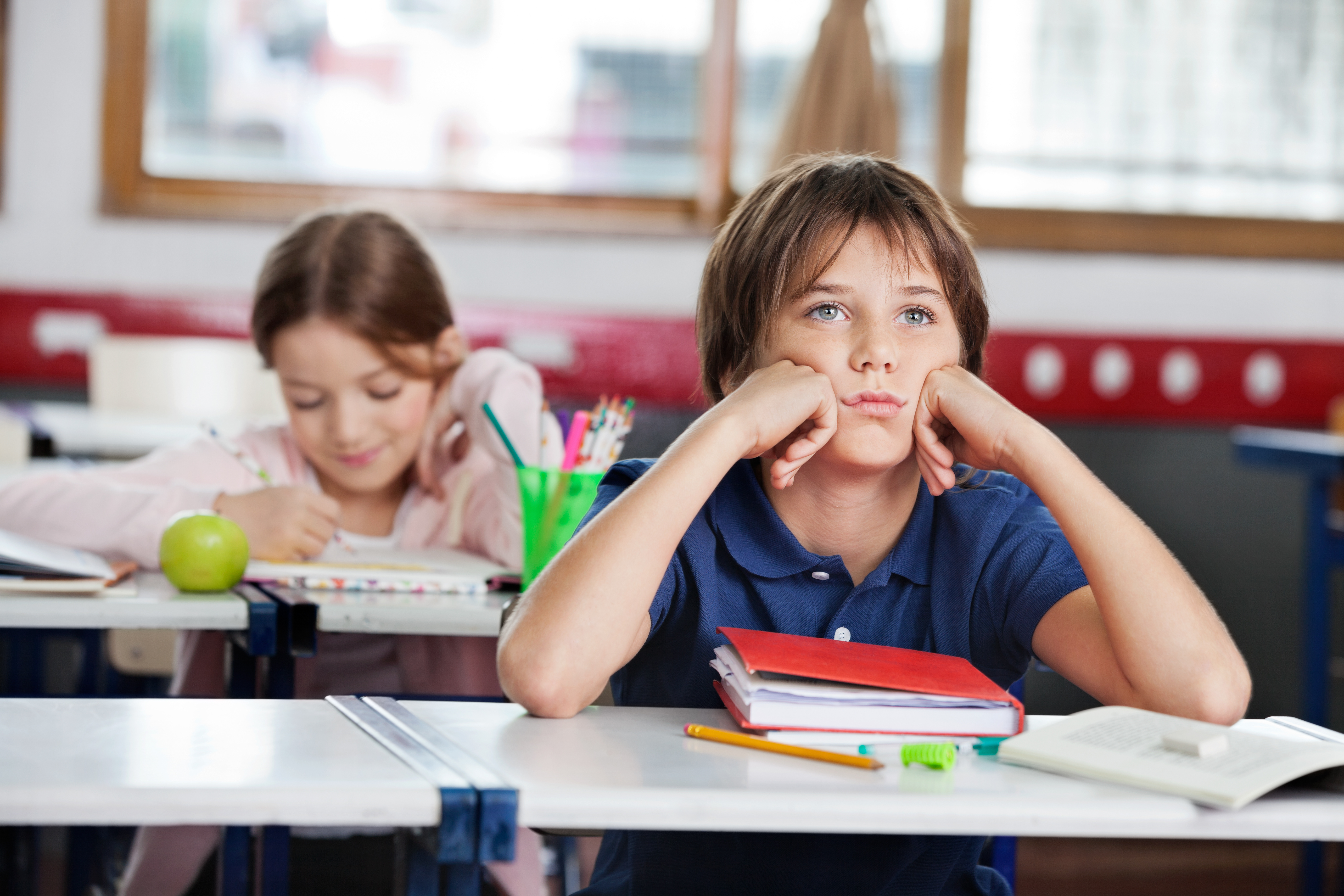 Bored schoolboy looking away while sitting at desk with girl in background at classroom