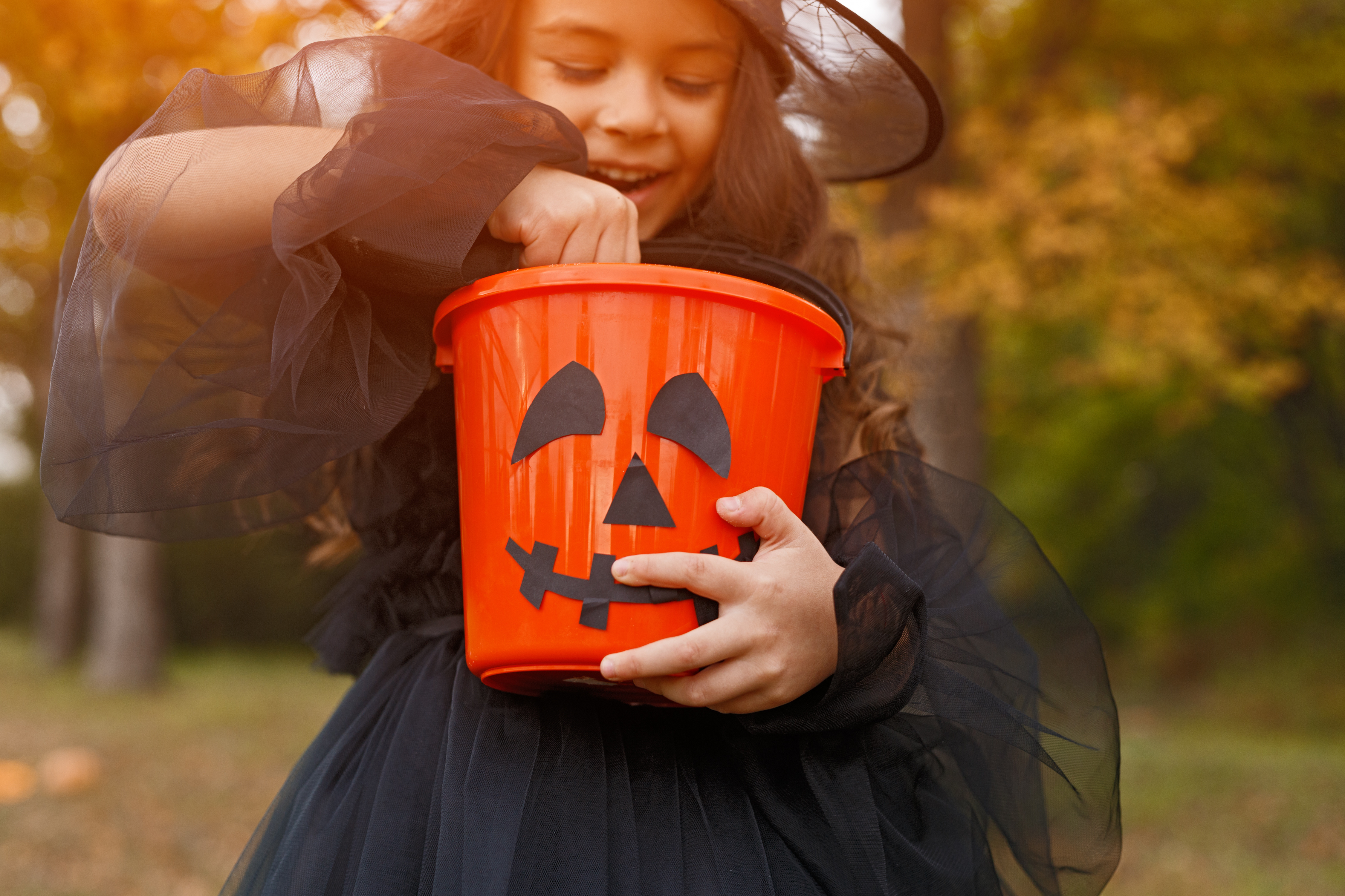 Crop happy girl in witch costume taking sweet from bucket while trick or treating in autumn park during Halloween celebration