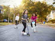 Three girls riding down the street on scooters and a bike