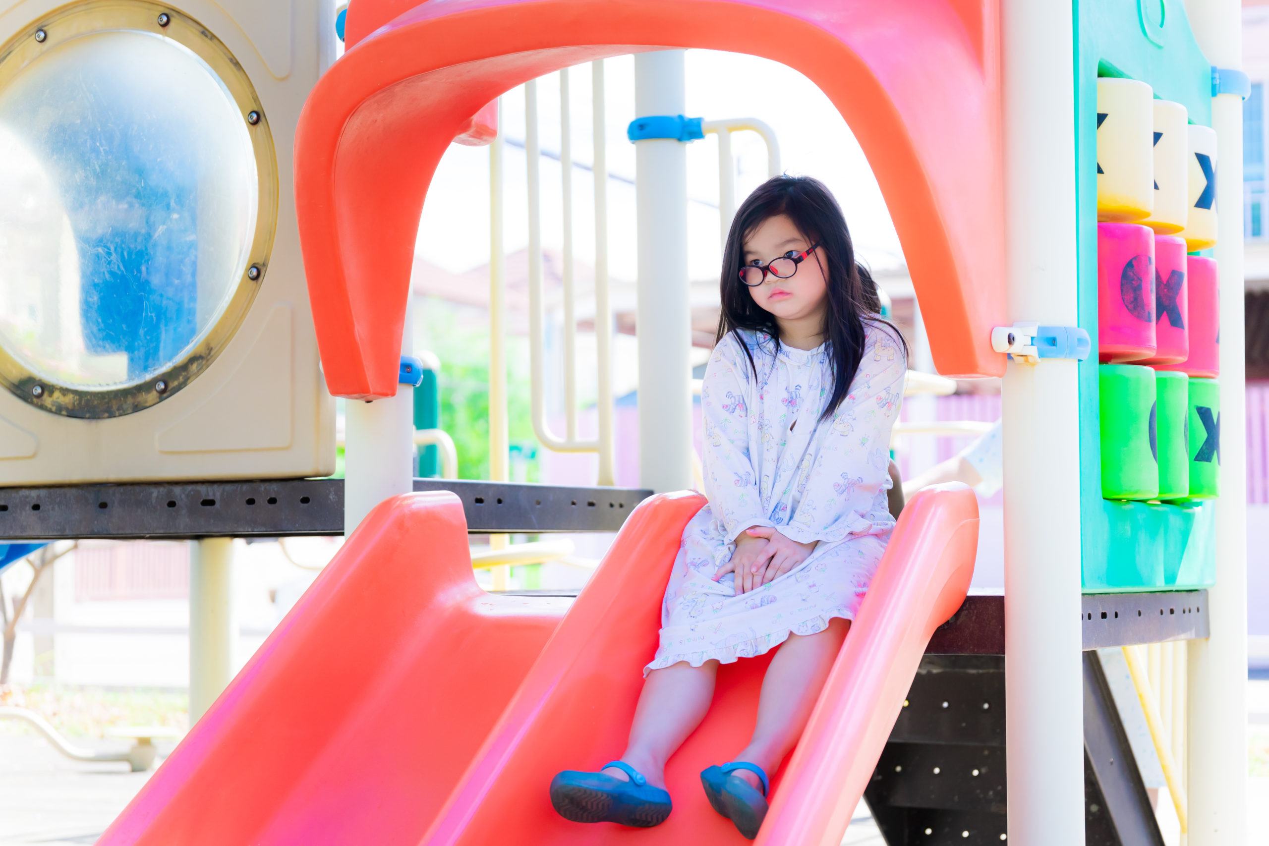 Asian child girl wearing glasses was bored at slider. Children in pajamas is sitting lonely at playground. Cute kid girl aged 6 years old.